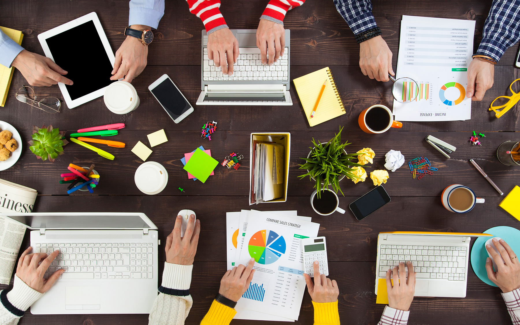 The hands of a group of business people working on various devices at an office desk.